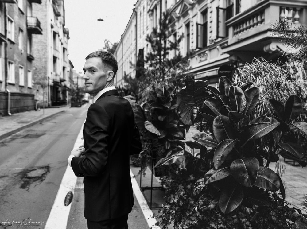 A groom in a black suit poses on an urban street with classic architecture.