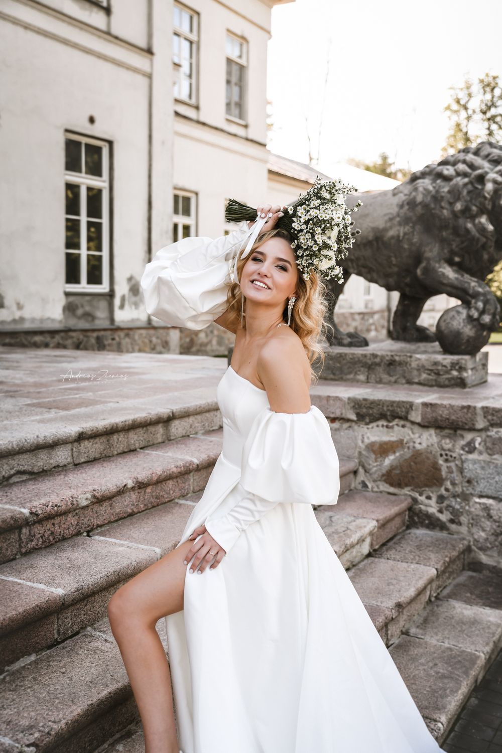 Bride in elegant off-shoulder white dress poses on stone steps outside historic building.