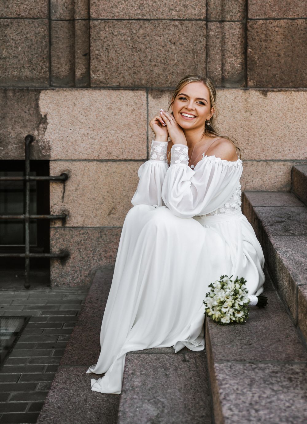 A bride in an off-shoulder gown sits elegantly on stone steps holding a white floral bouquet.