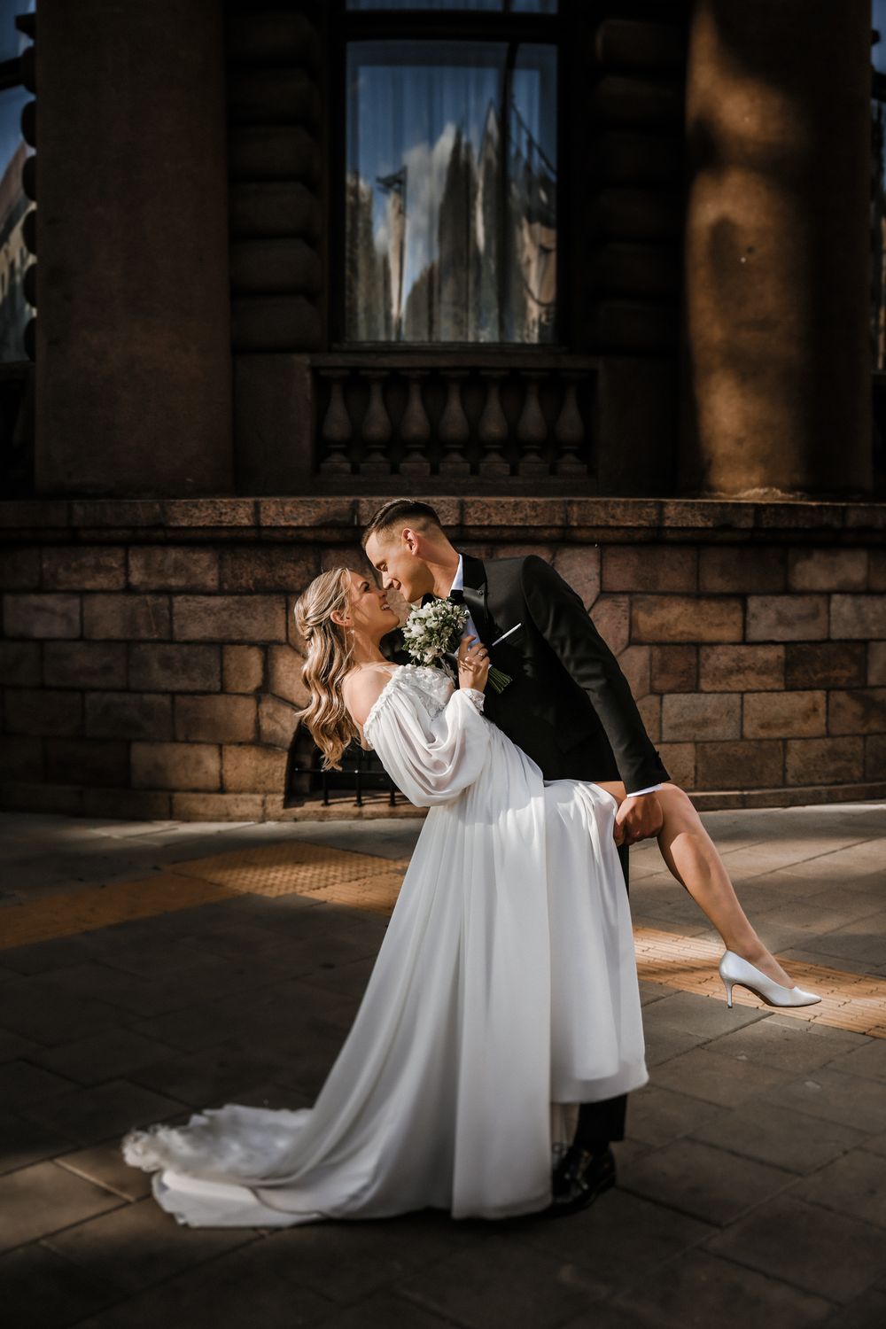 Dramatic dip kiss between newlyweds against stone building architecture.