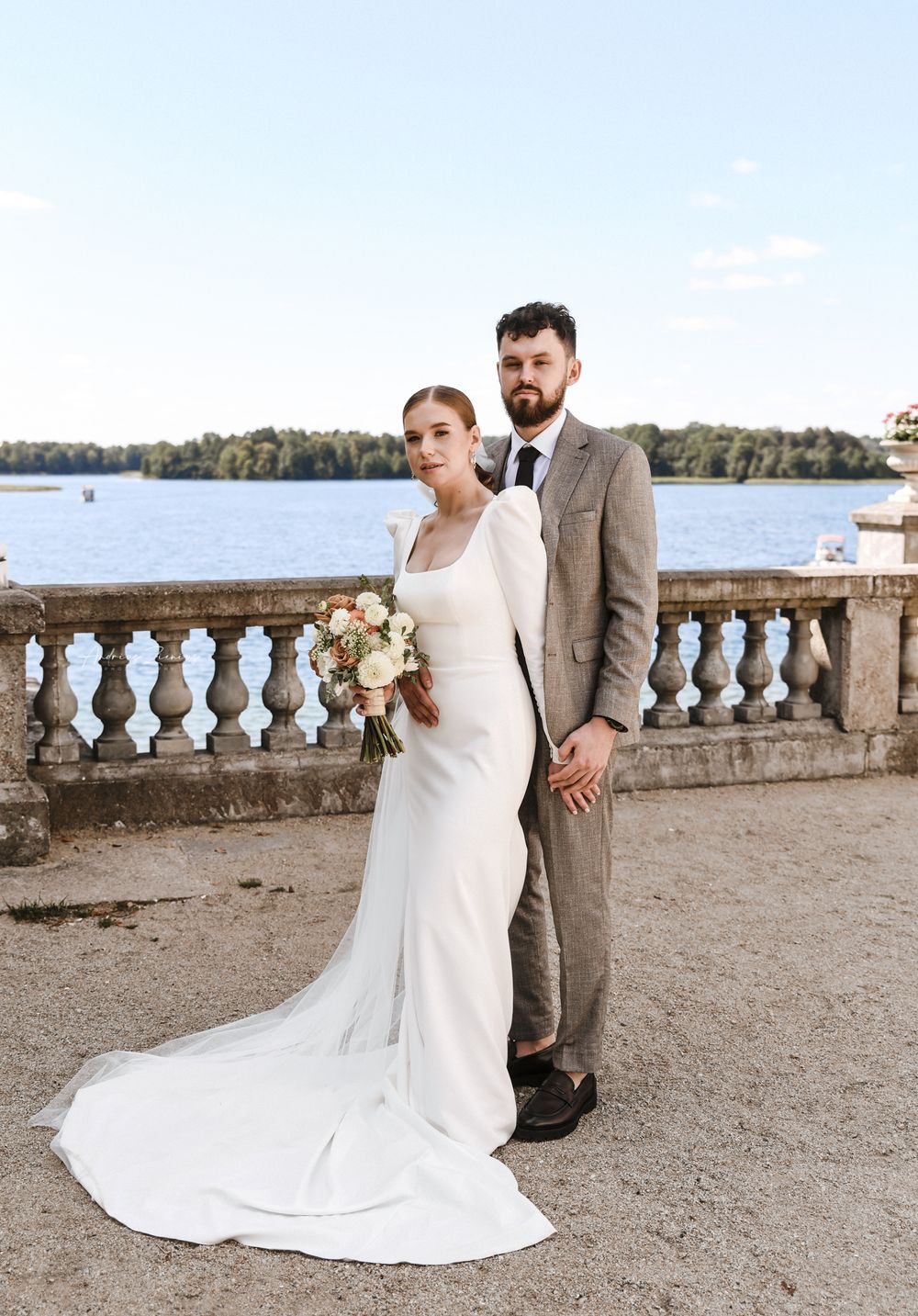 A bride and groom pose against a waterfront balustrade with scenic lake views in the background.