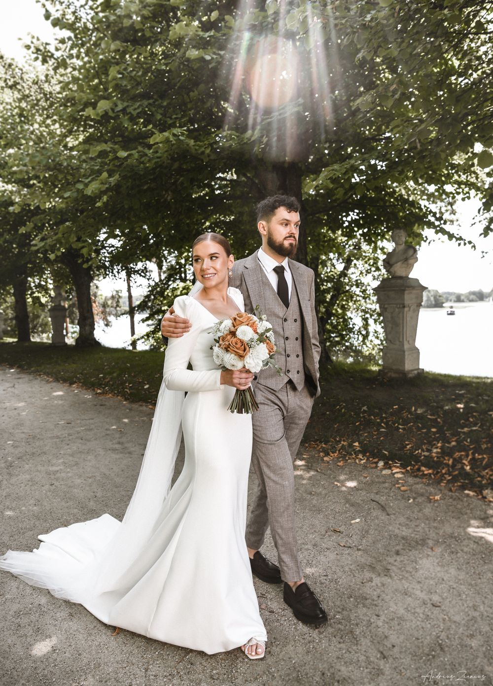 Newlyweds walking along a tree-lined path by a lake, bride holding autumn-colored bouquet.