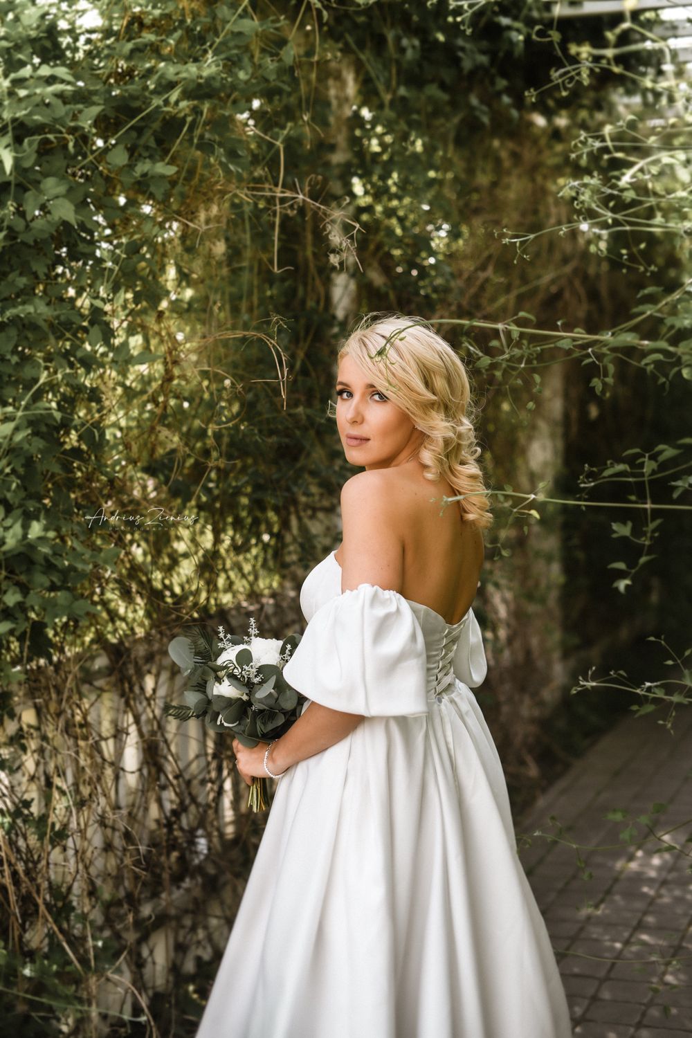 A bride in an off-shoulder white wedding dress poses against a garden backdrop with sunlit foliage.