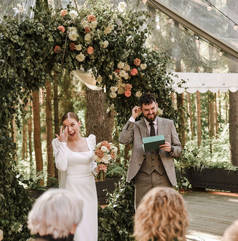 A couple shares a laugh while reading vows under a floral arch during their outdoor forest ceremony.