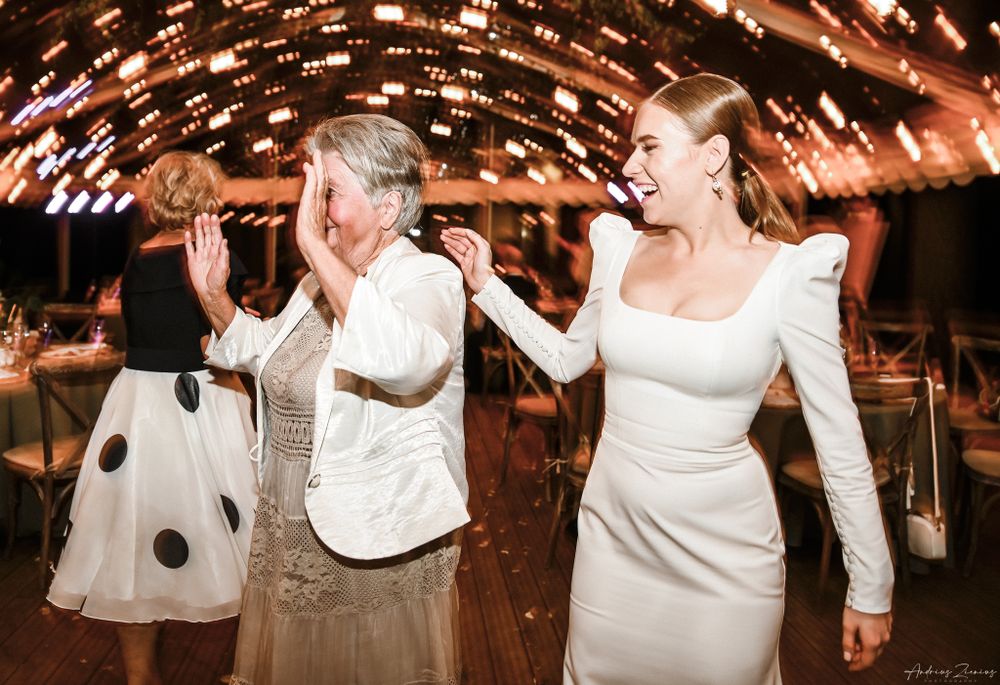 Two women in white dresses dancing joyfully under sparkling reception lights.