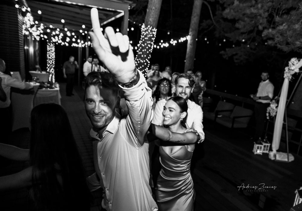 A lively black and white photo of wedding guests dancing under string lights at an outdoor reception.