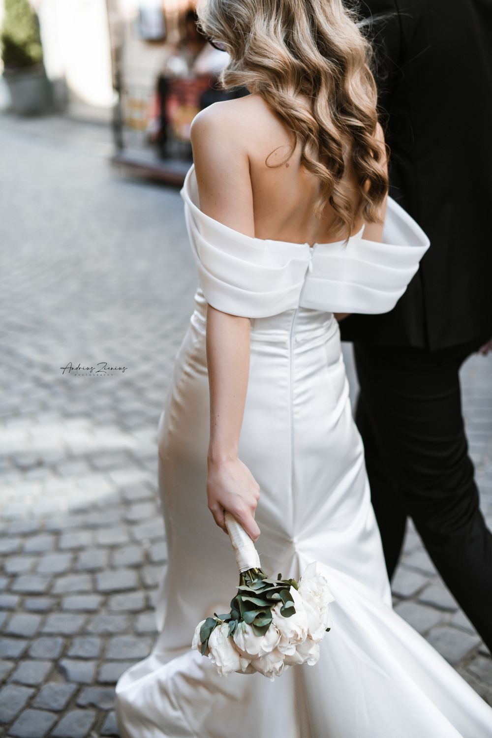 Back view of an elegant white wedding dress with off-shoulder design on cobblestone street.