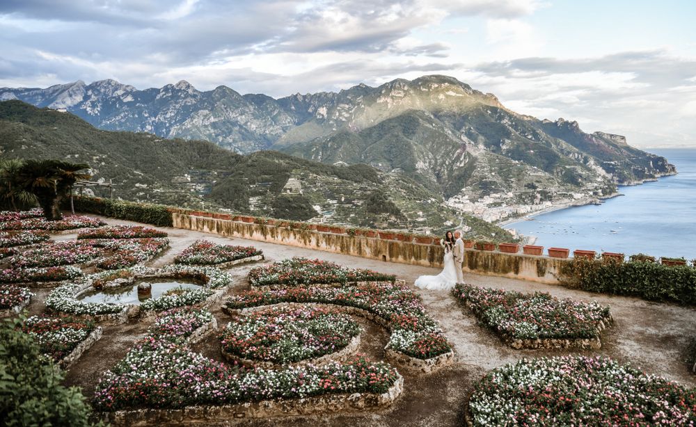 Aerial view of ornate garden terraces overlooking the Amalfi Coast with mountains and sea in the background.