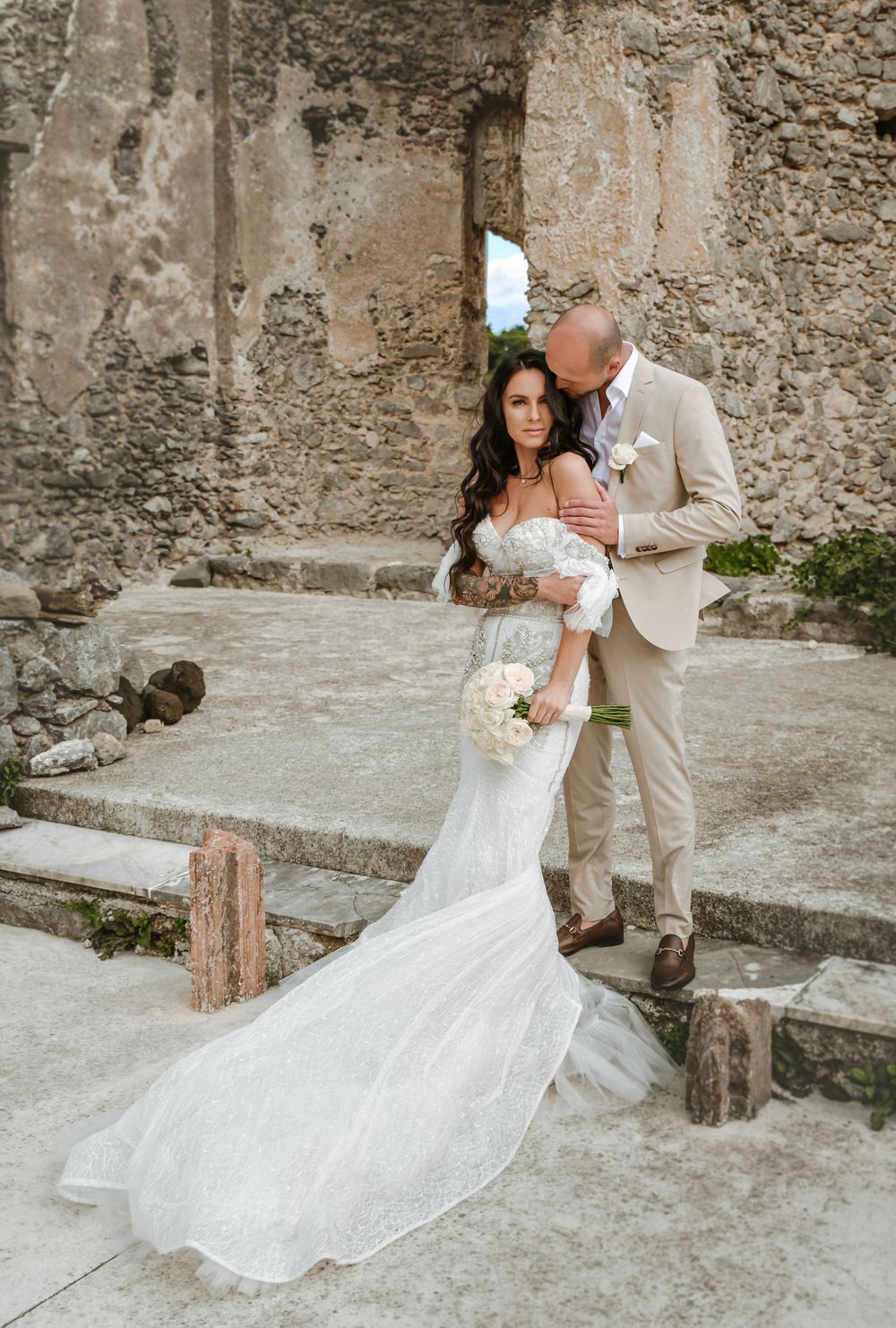 A romantic wedding photo against ancient stone walls showcasing a flowing white lace gown.