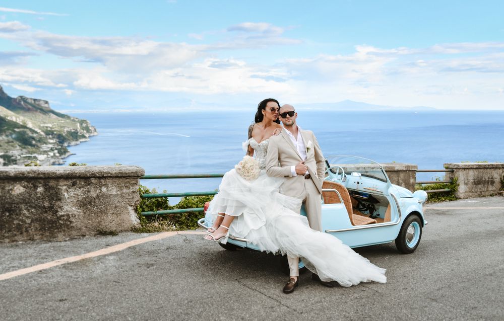 Wedding couple poses in front of classic car at scenic coastal location for romantic photoshoot.
