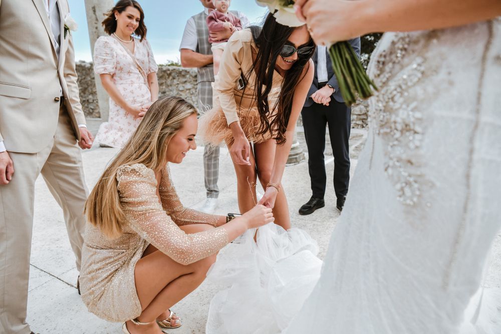 Wedding party members help adjust the bride's dress on a sunny terrace.