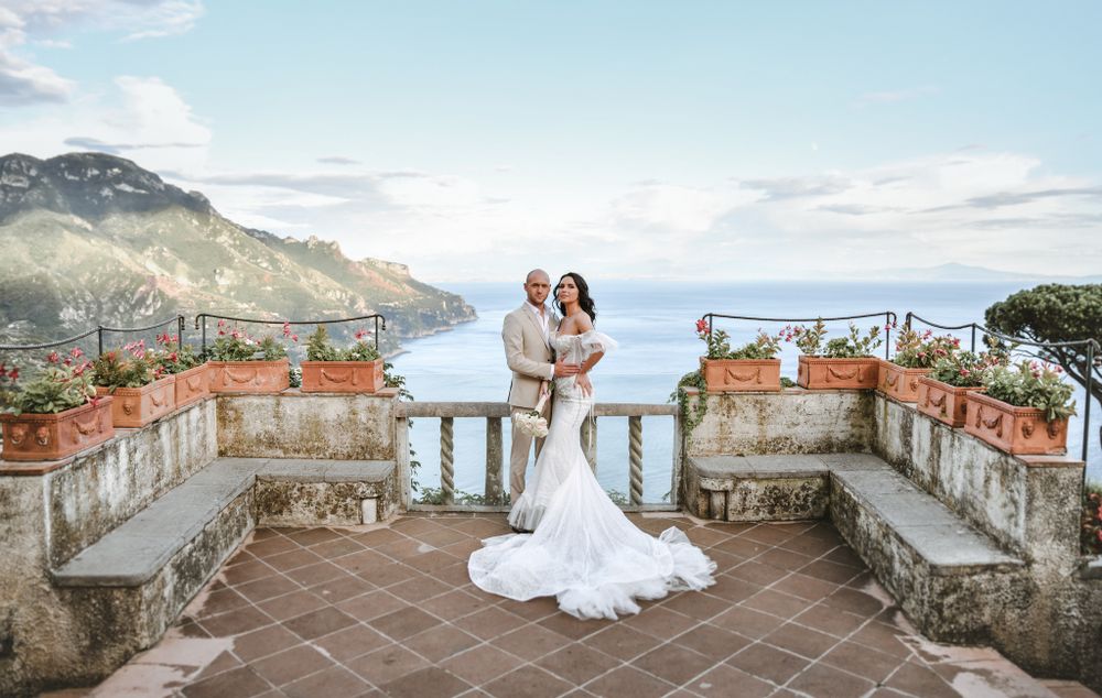 A wedding couple poses on a historic terrace overlooking the Mediterranean coastline at sunset.