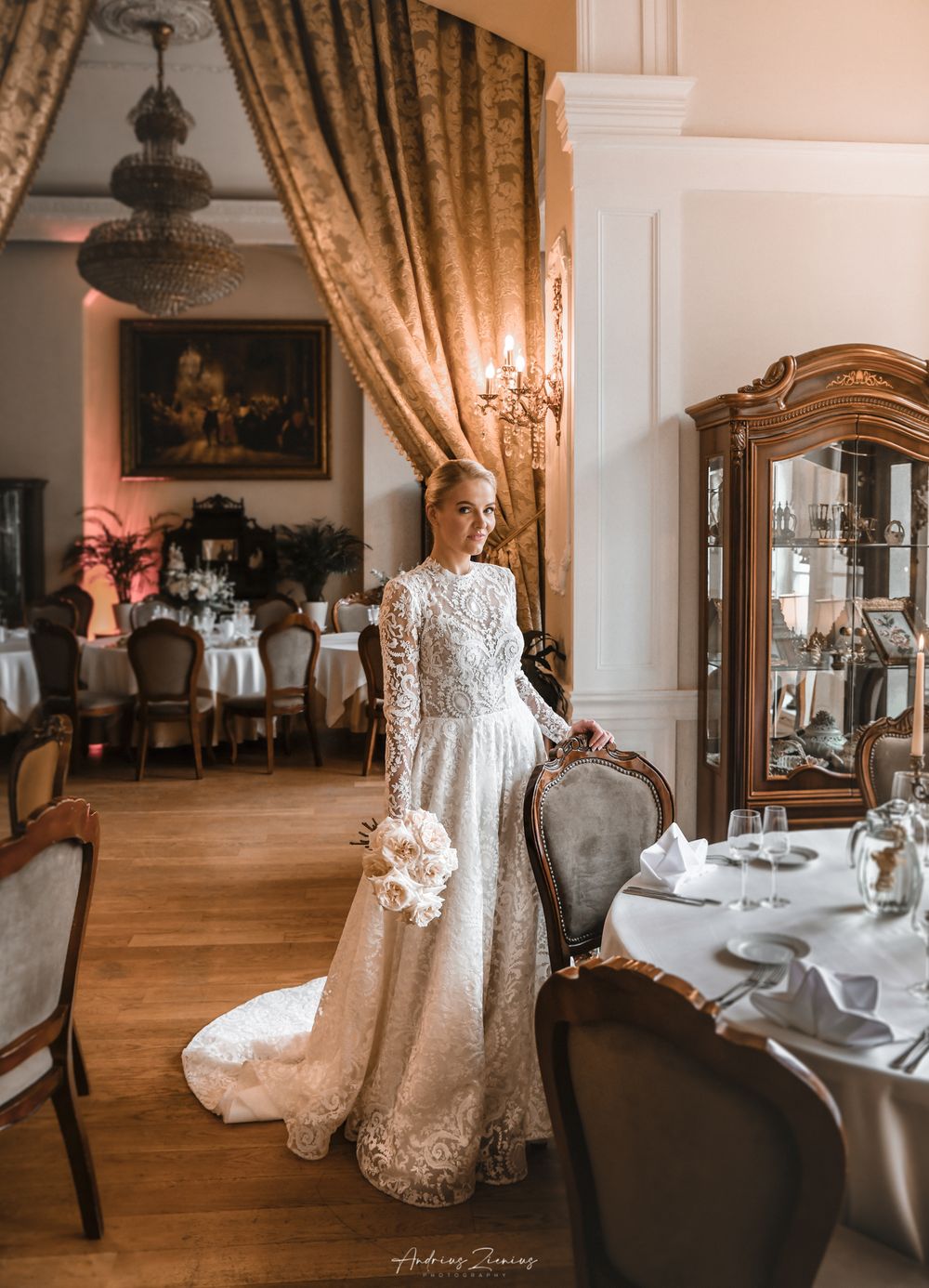 Elegant bride in lace gown poses in luxurious dining room with chandelier and golden curtains.