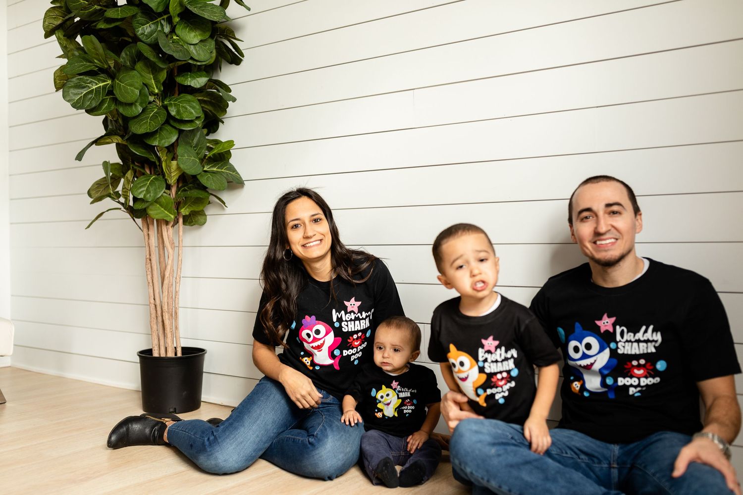 Family wearing matching black t-shirts sitting together against a white wall with a plant.