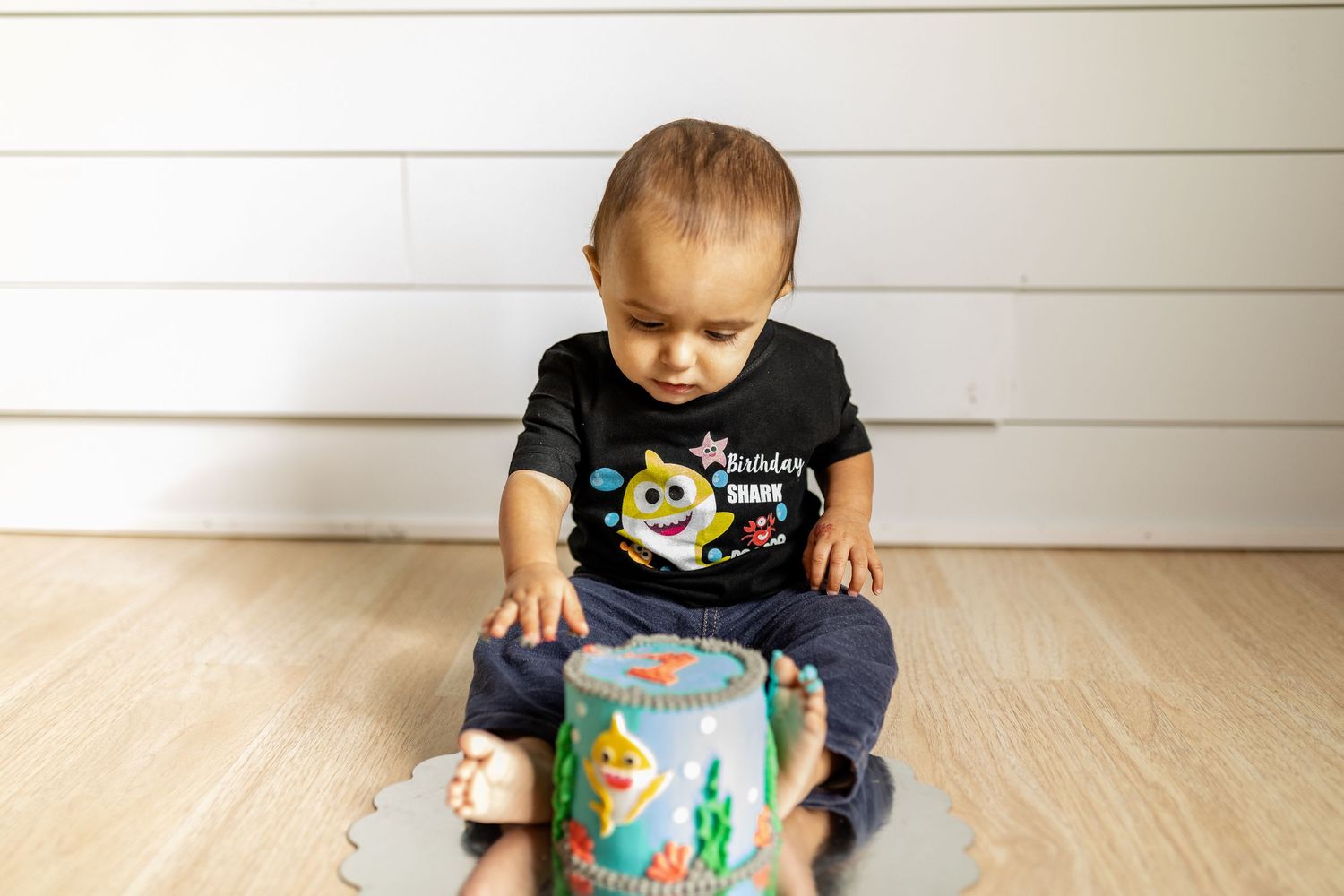 Young child in black t-shirt playing with a teal decorated container on wooden floor.