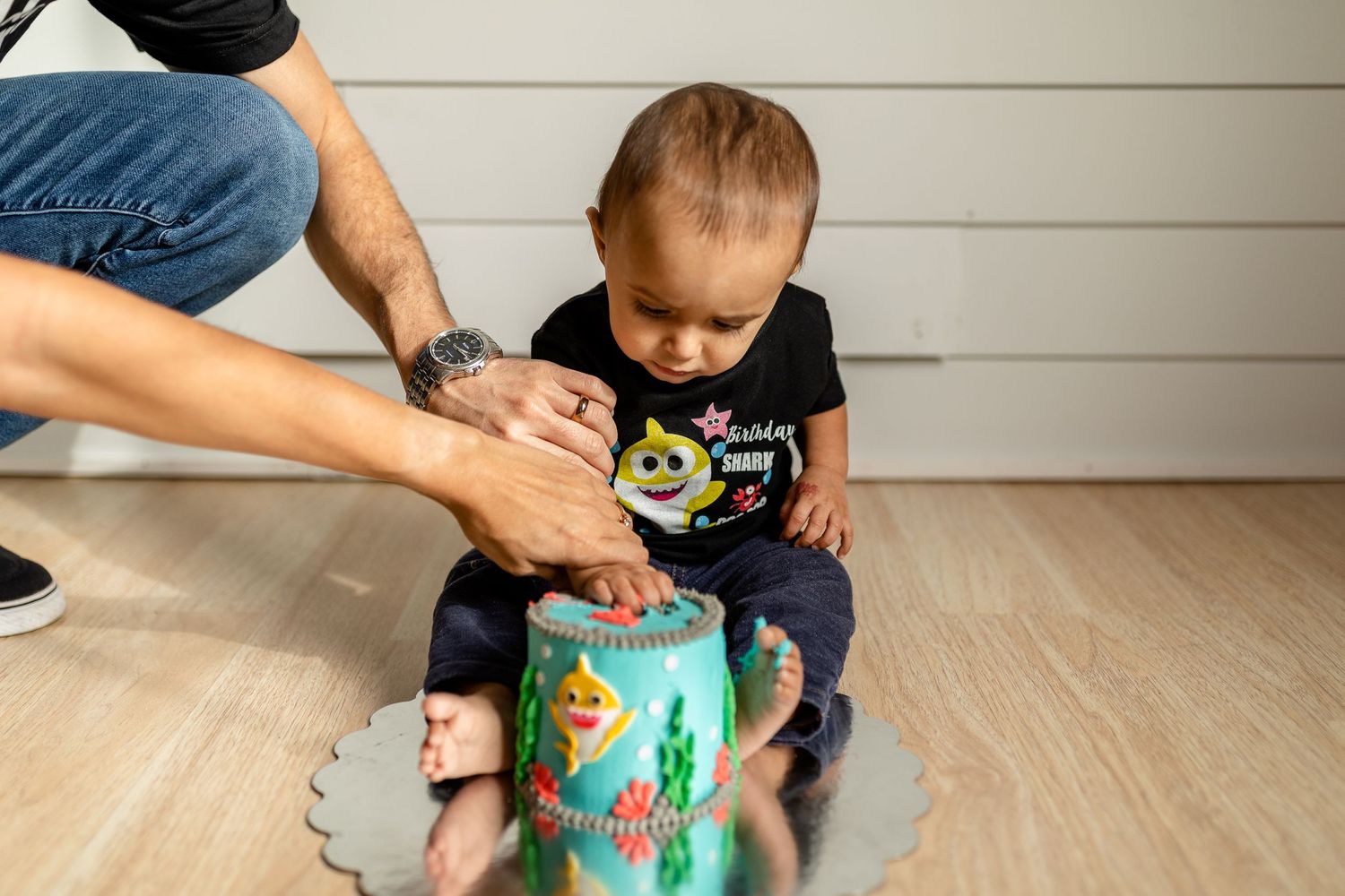 Adult hands helping small child with decorated teal container on wooden floor.