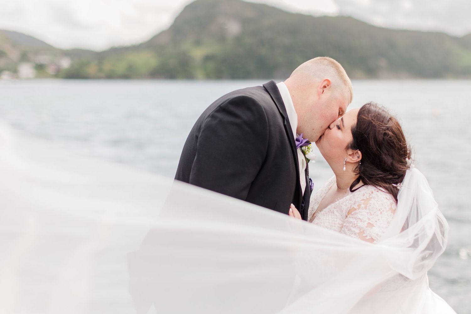 Romantic seaside wedding portrait with flowing veil and mountain backdrop.