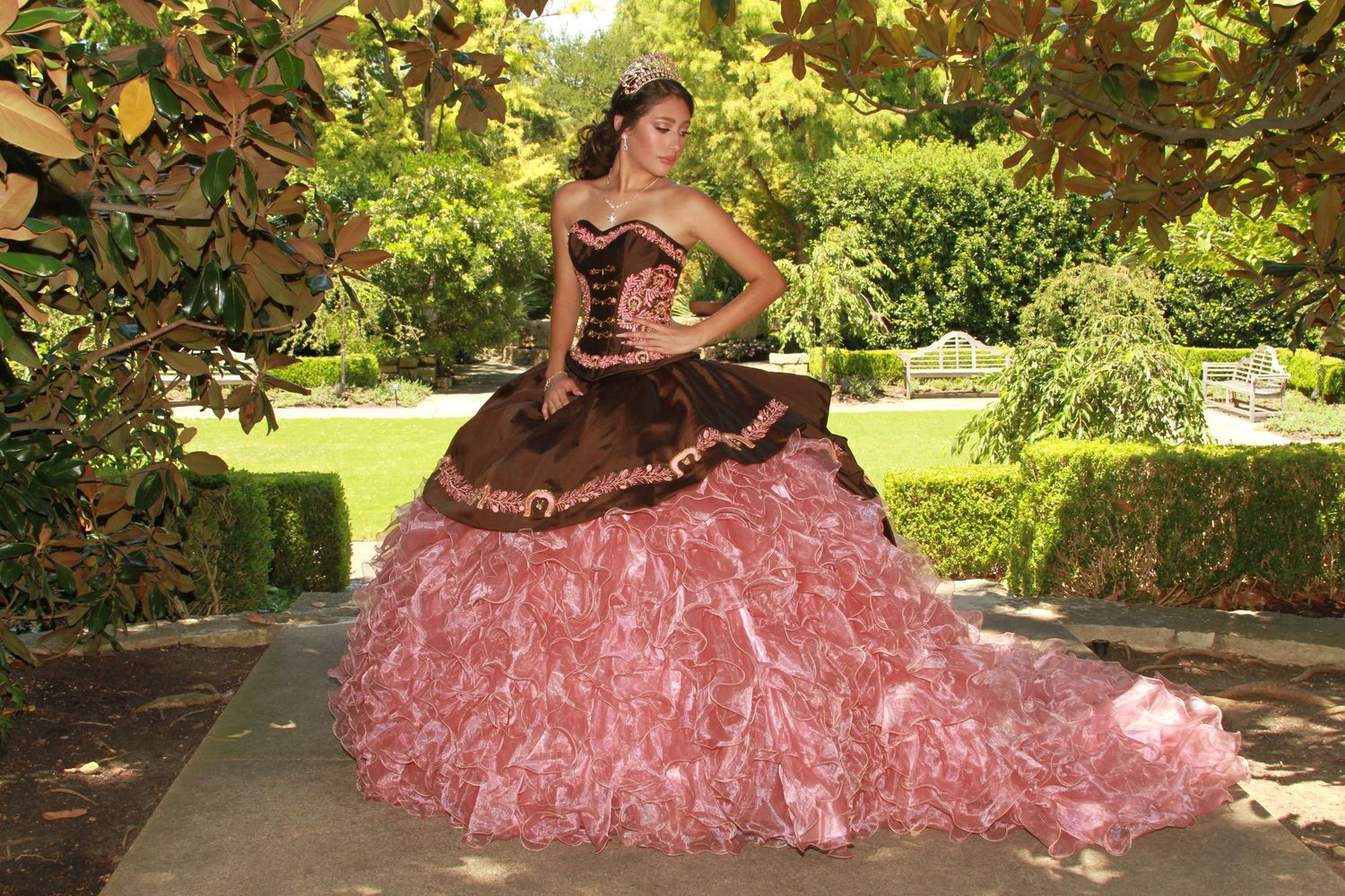 A coral pink quinceañera dress photographed in a lush garden setting.