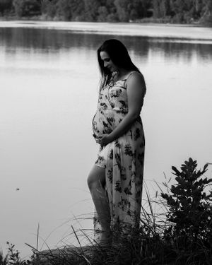 Silhouette of someone in a dress standing by a tranquil lake with vegetation in the foreground.