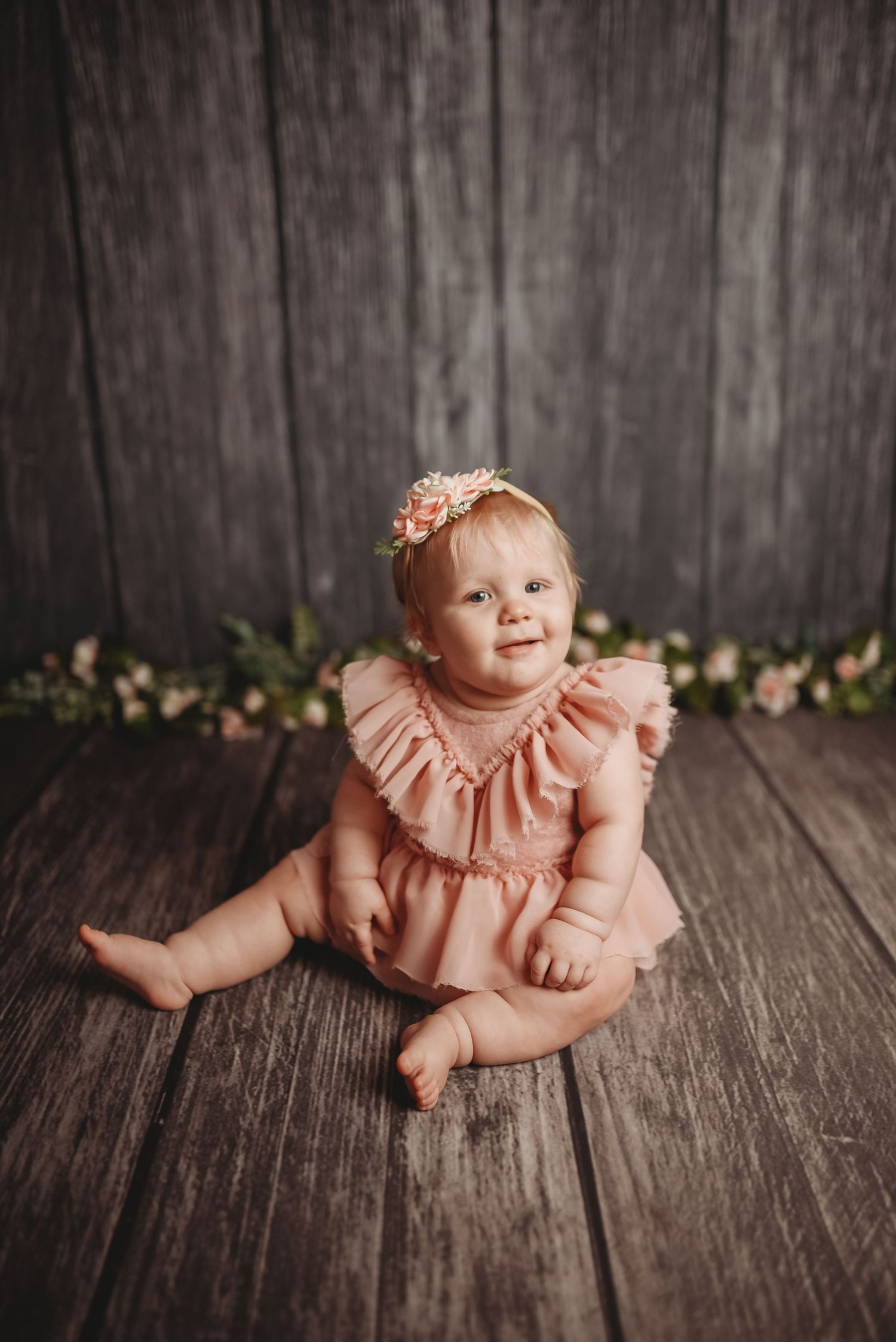 Baby in pink ruffled dress sits on dark wooden floor with floral accents.