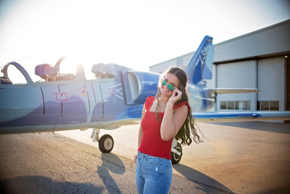 Young woman in red top and jeans wearing sunglasses posing near a blue airplane at an airfield.