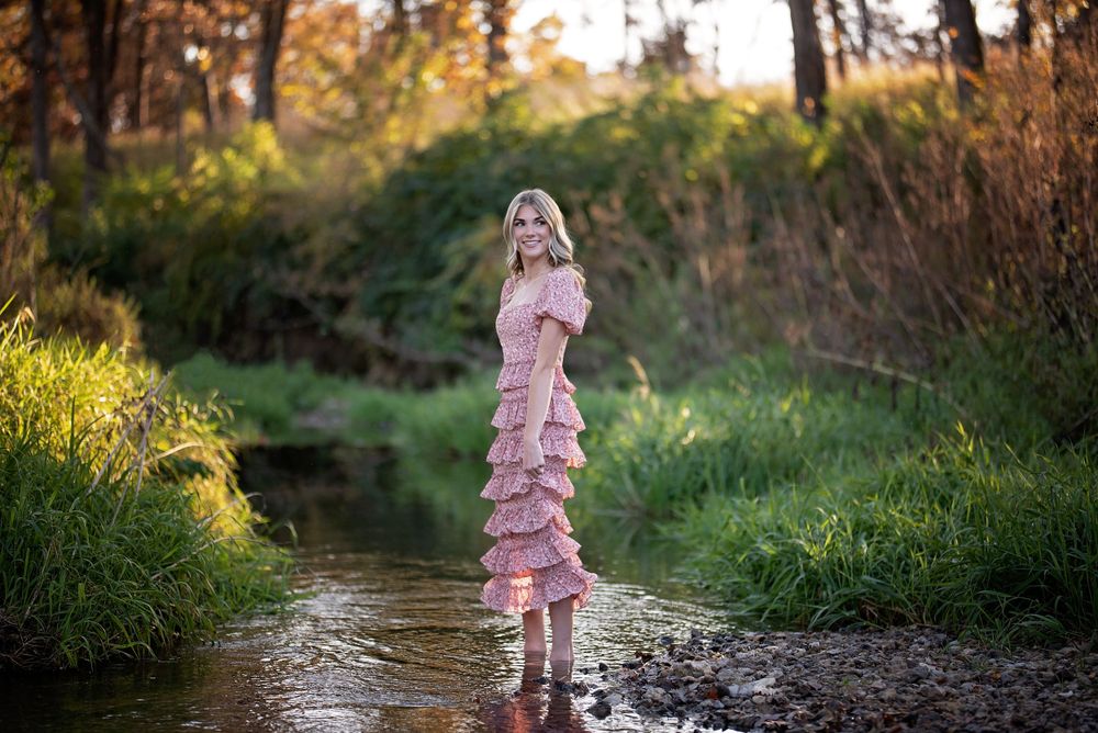 Young woman in a pink ruffled dress standing in a shallow creek surrounded by autumn foliage.
