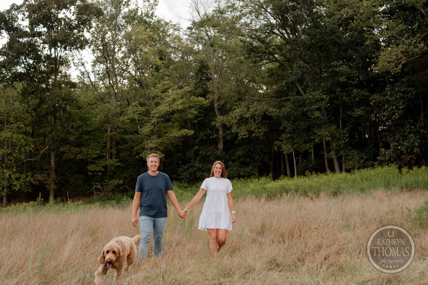 A couple walking through a golden field with their goldendoodle dog at sunset near the woods.