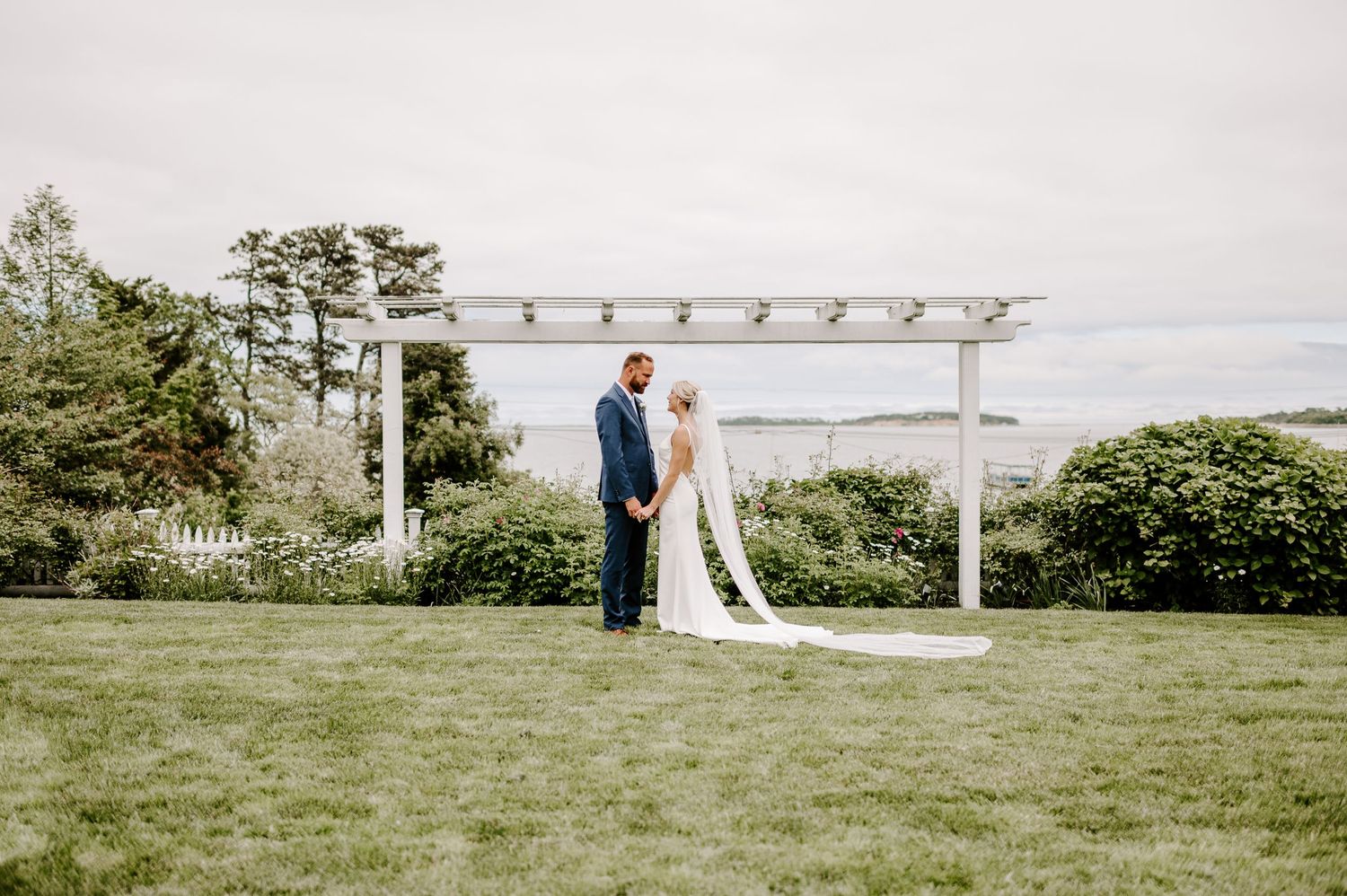 A serene wedding scene with couple standing under white pergola overlooking coastal landscape.