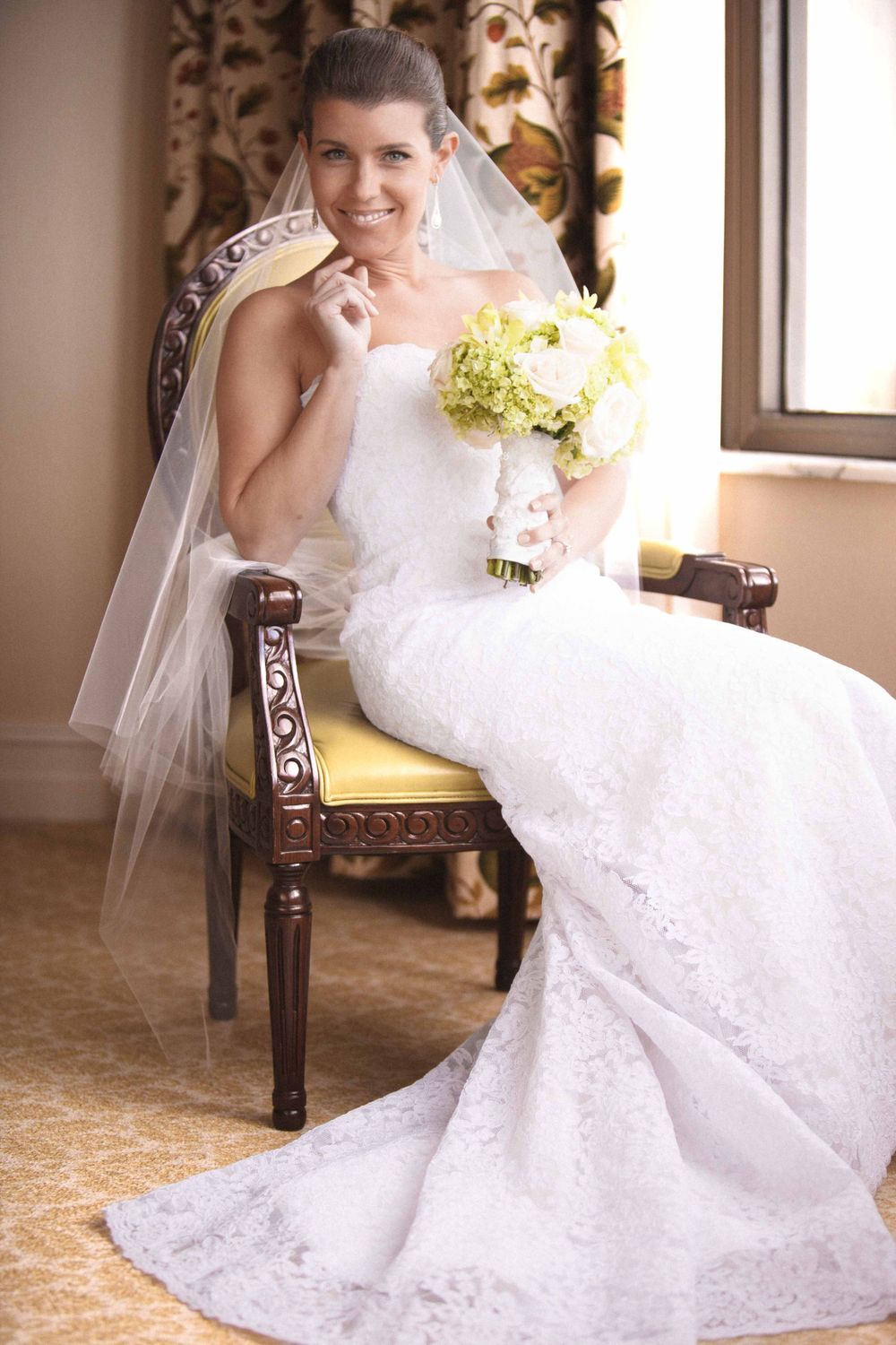 A bride in a white lace wedding gown and veil poses elegantly on an upholstered chair in an ornate indoor setting.