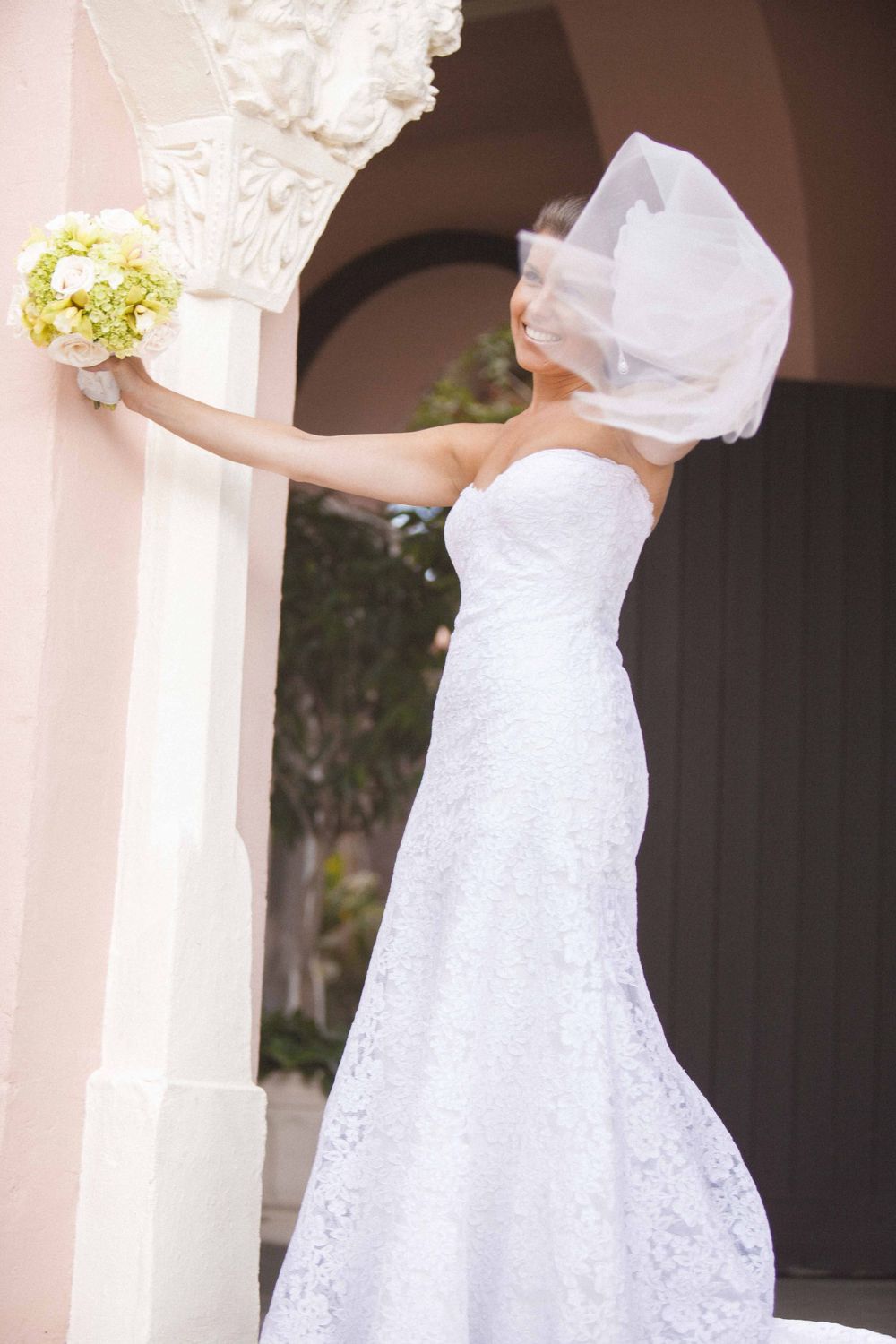 A bride in an elegant white lace wedding gown and veil holds up a yellow bouquet against a light pink wall.