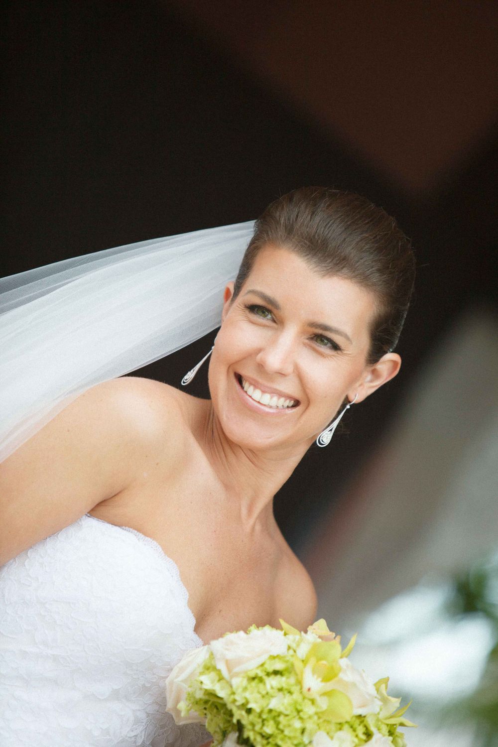 A bride in a white strapless wedding gown holds a green and white bouquet while smiling brightly.