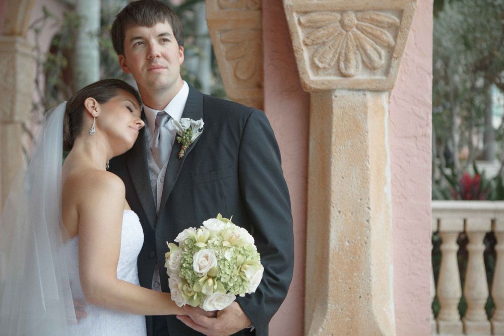 A romantic wedding couple stands by an ornate stone column, with the bride holding a white hydrangea bouquet.