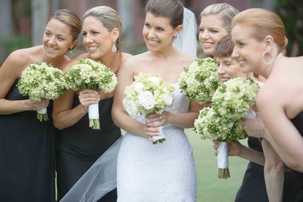 A wedding party smiles and laughs together while holding green and white hydrangea bouquets outdoors.