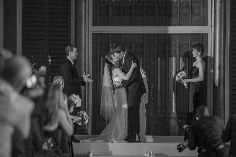 A black and white photograph captures an intimate moment during a wedding ceremony in a grand indoor venue.