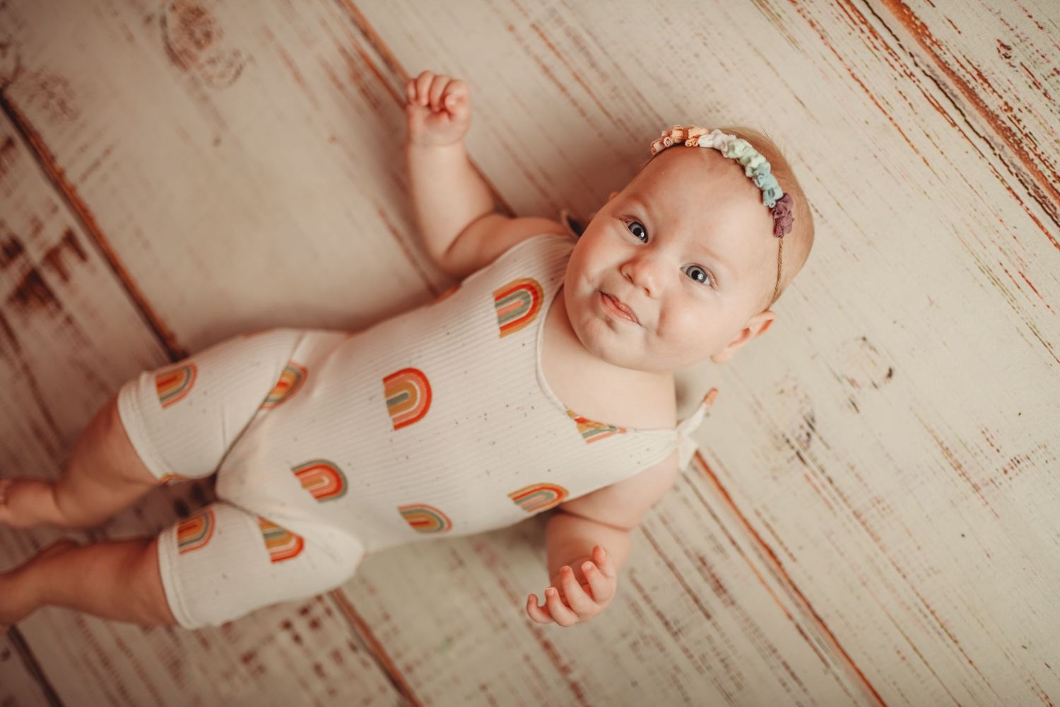 Baby in white outfit lies on distressed wooden floor during photo session.
