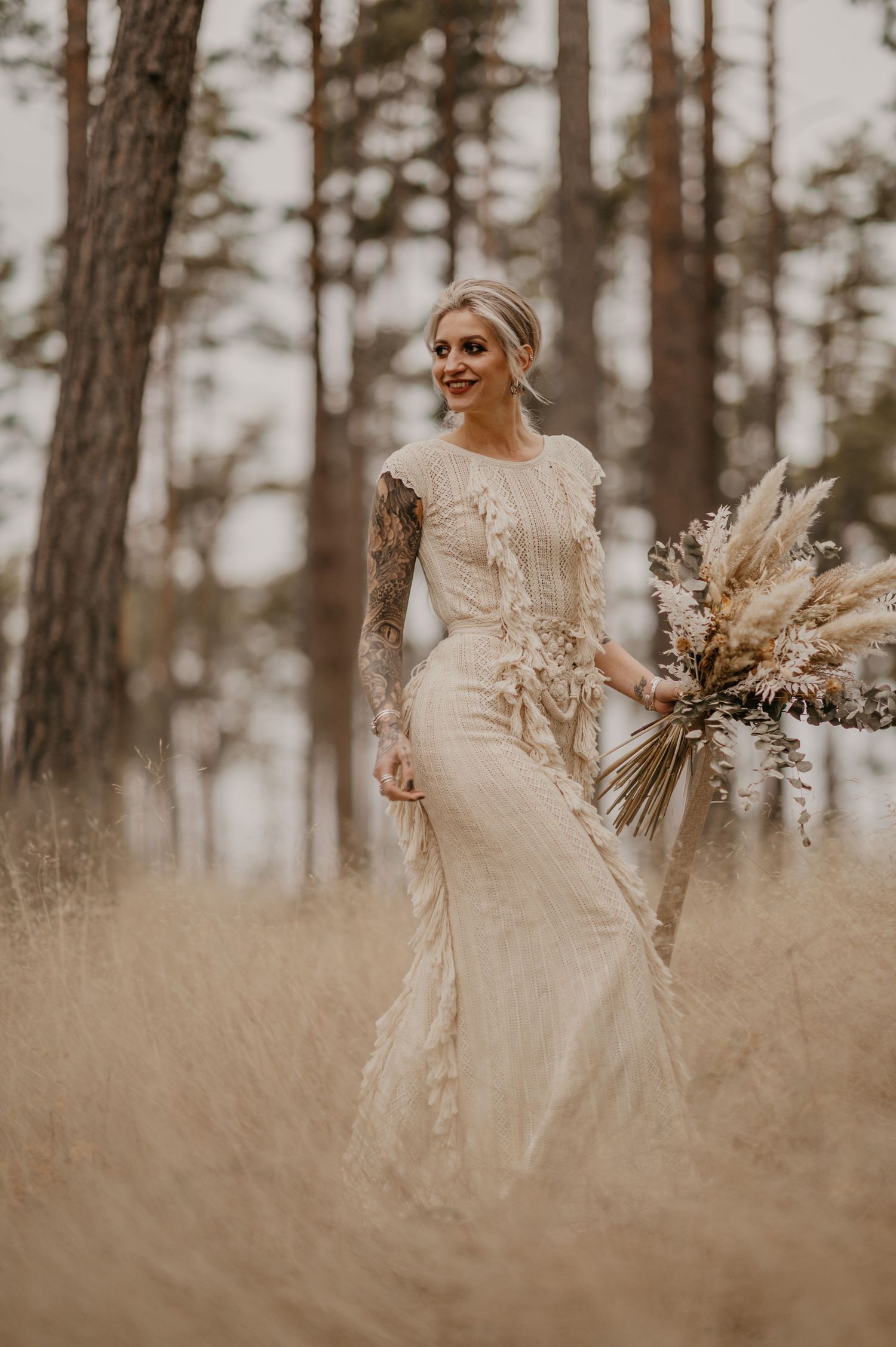 Bride in lace long-sleeve wedding gown holding pampas grass bouquet, standing in golden field surrounded by pine trees.
