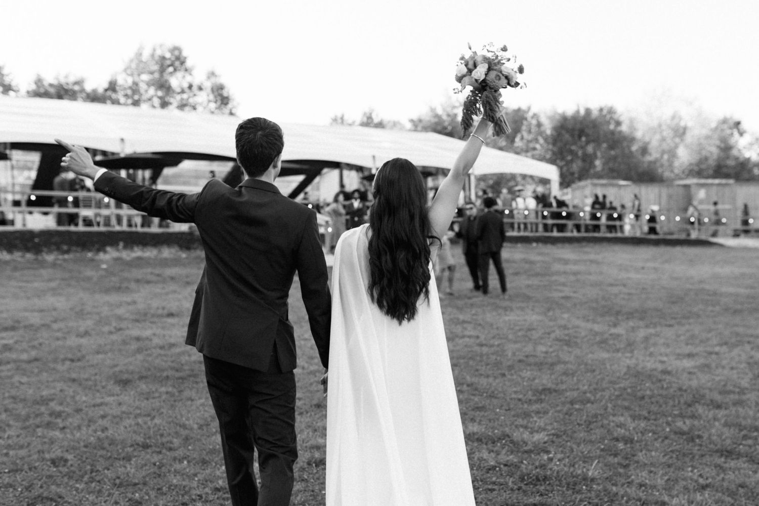 Bride and groom hold hands, raising their other arms up in celebration as they walk toward their reception tent.