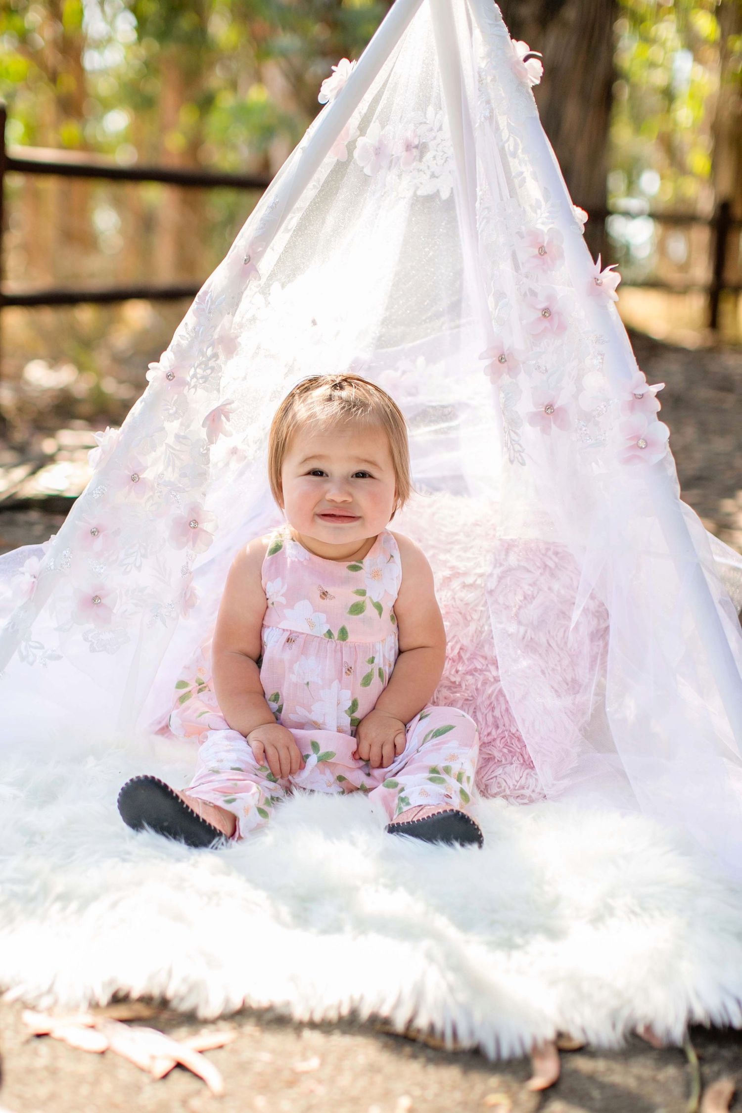 Small child sitting on white fur inside delicate white teepee tent decorated with pink flowers in outdoor setting.