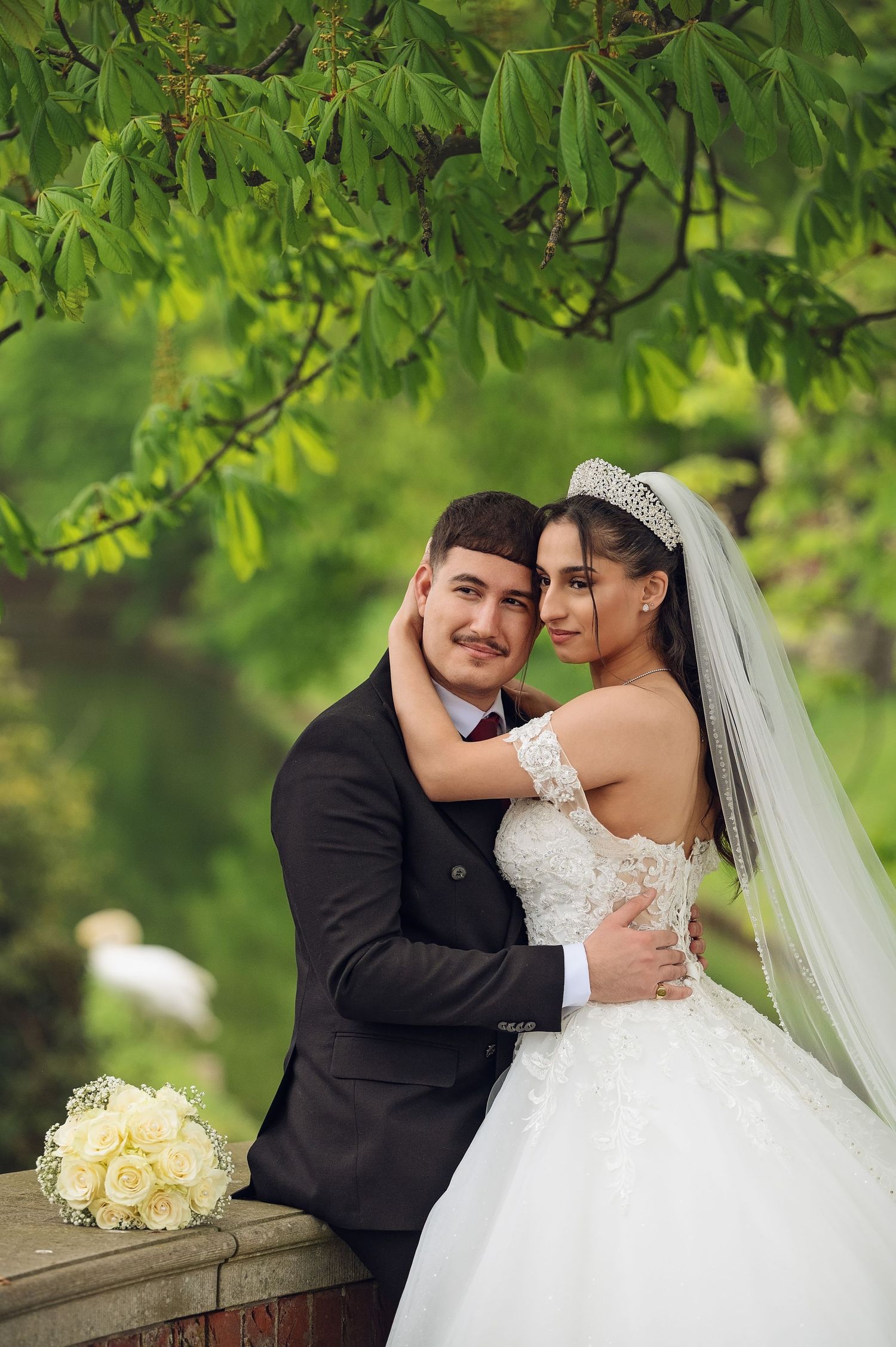 Wedding couple in formal attire embracing under green tree canopy with white bouquet.