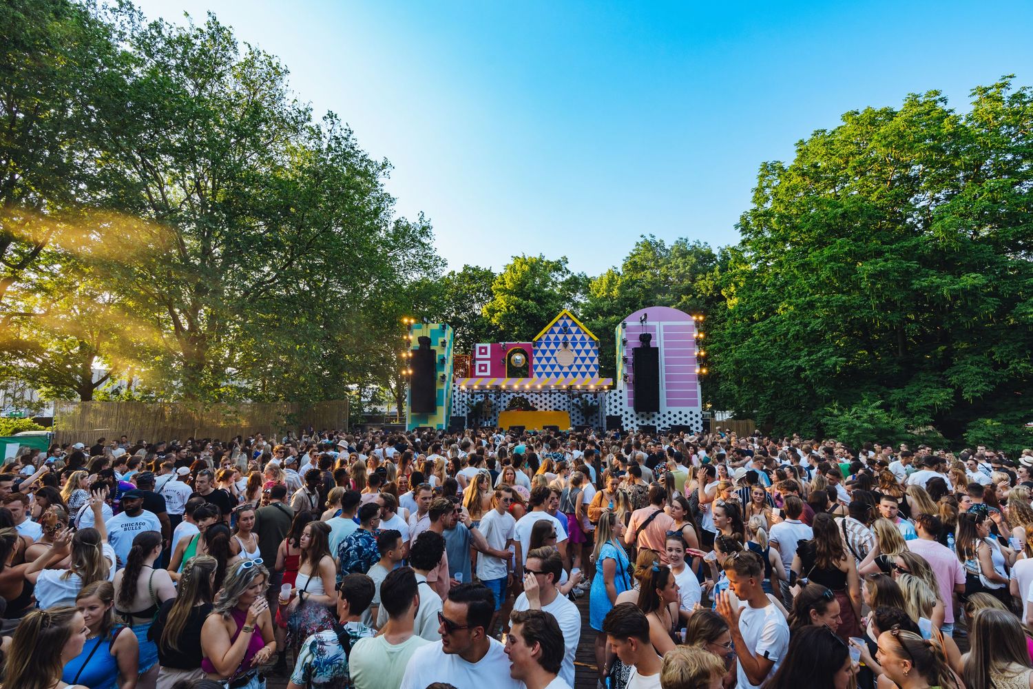 Large outdoor music festival crowd gathered in front of a stage on a sunny summer day.