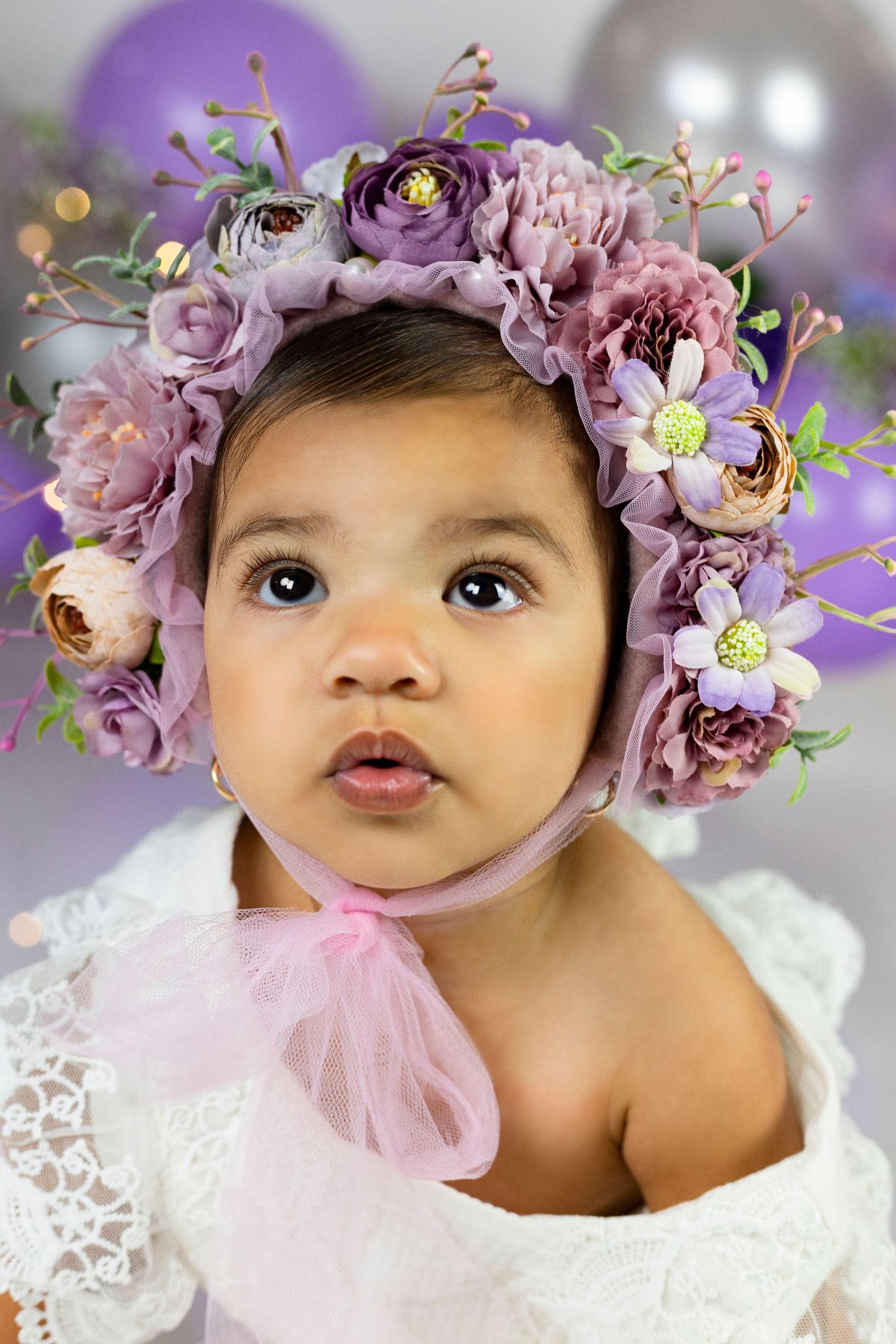 Baby wearing purple floral crown and white lace dress with balloons in background.