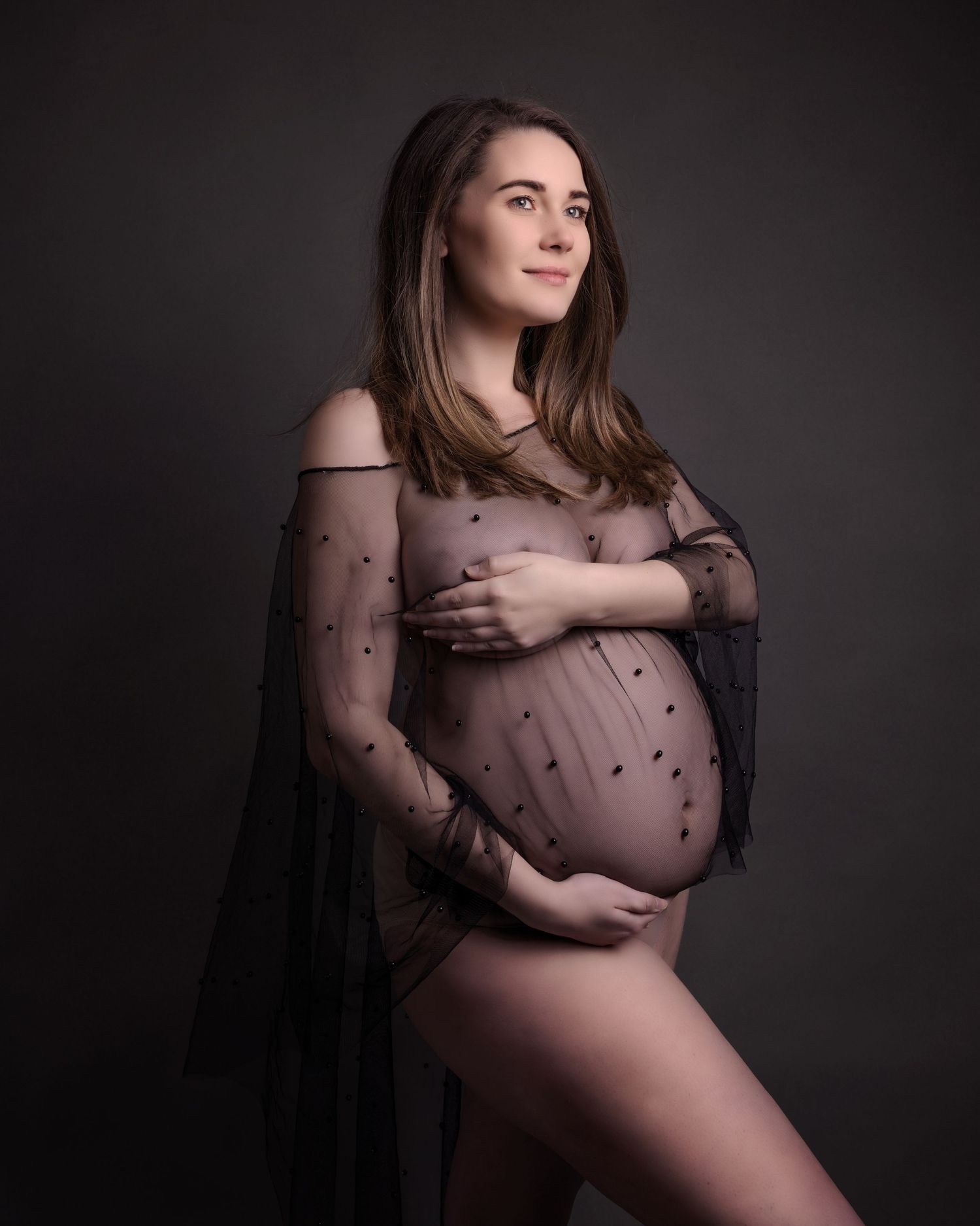 Artistic maternity portrait showing an elegant sheer black overlay dress against dark backdrop.