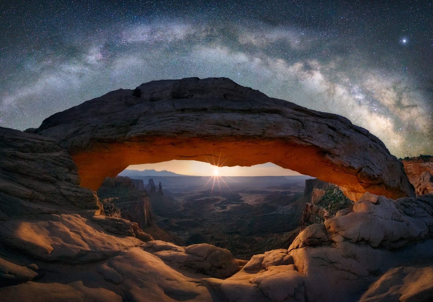 The Milky Way galaxy arches over Mesa Arch in Canyonlands National Park at night with glowing orange rock formations.