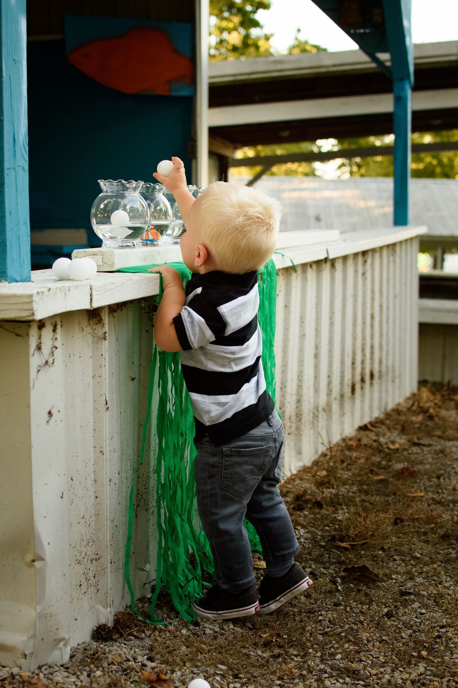 Curious toddler boy in striped shirt reaching up to explore a fishbowl at a carnival game booth.
