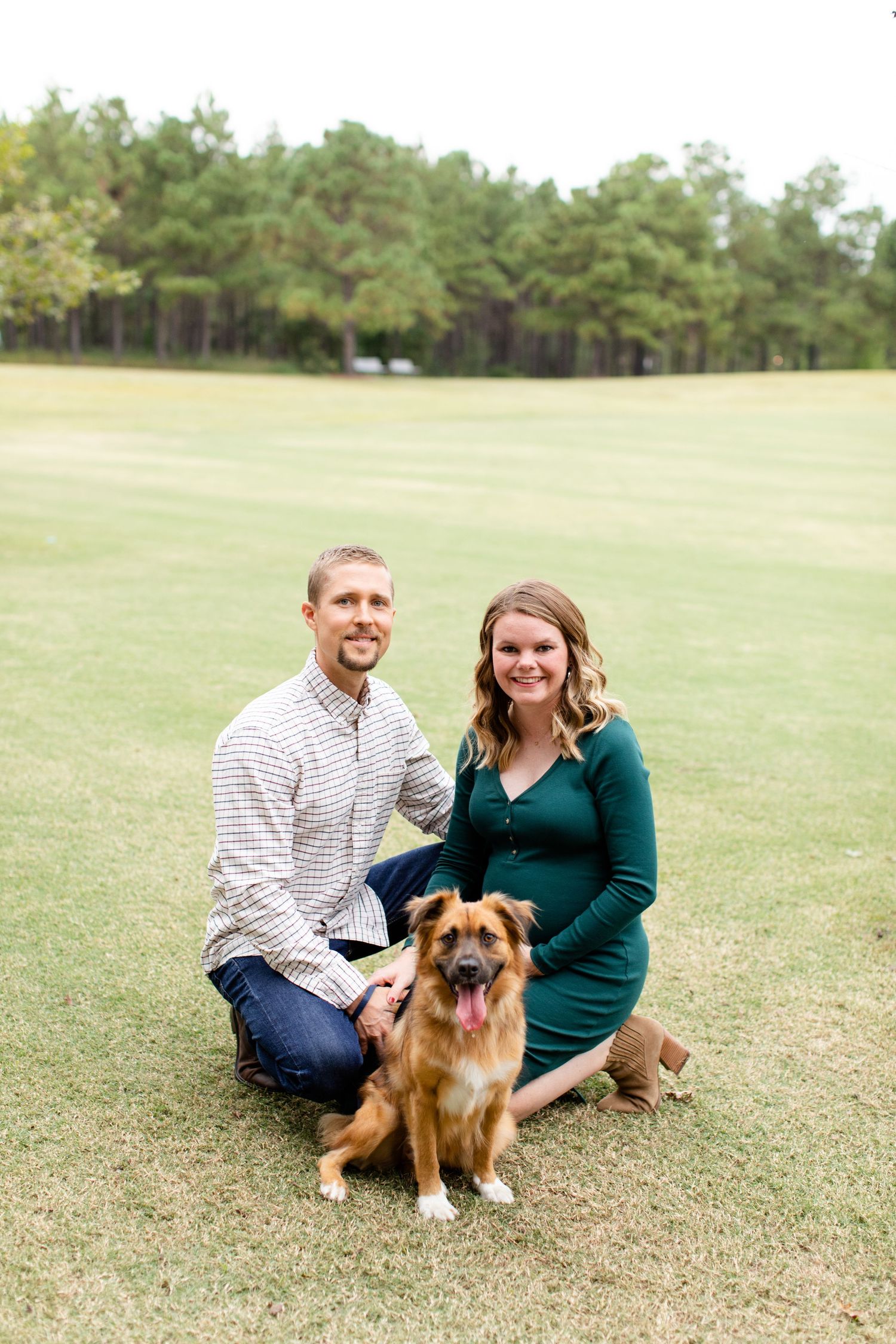 Couple sits with their German Shepherd mix dog on a grassy field surrounded by pine trees.