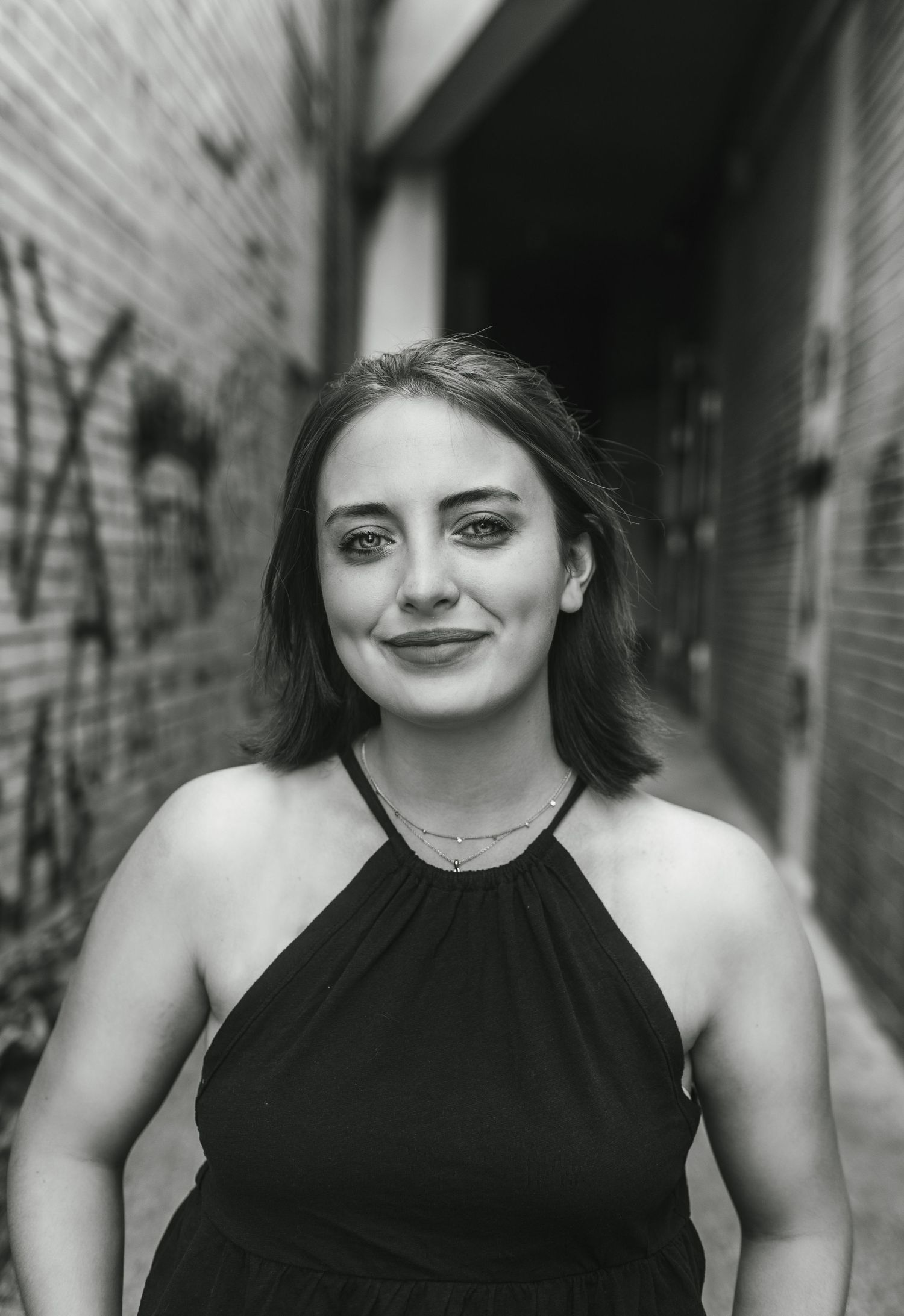A black and white portrait of a smiling woman wearing a halter top dress against an urban backdrop.