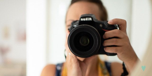 A photographer holds up a Nikon DSLR camera in a self-portrait style shot.
