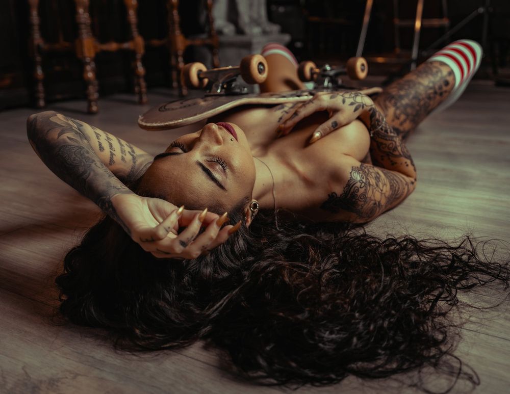 Moody indoor portrait with skateboard and intricate body art.