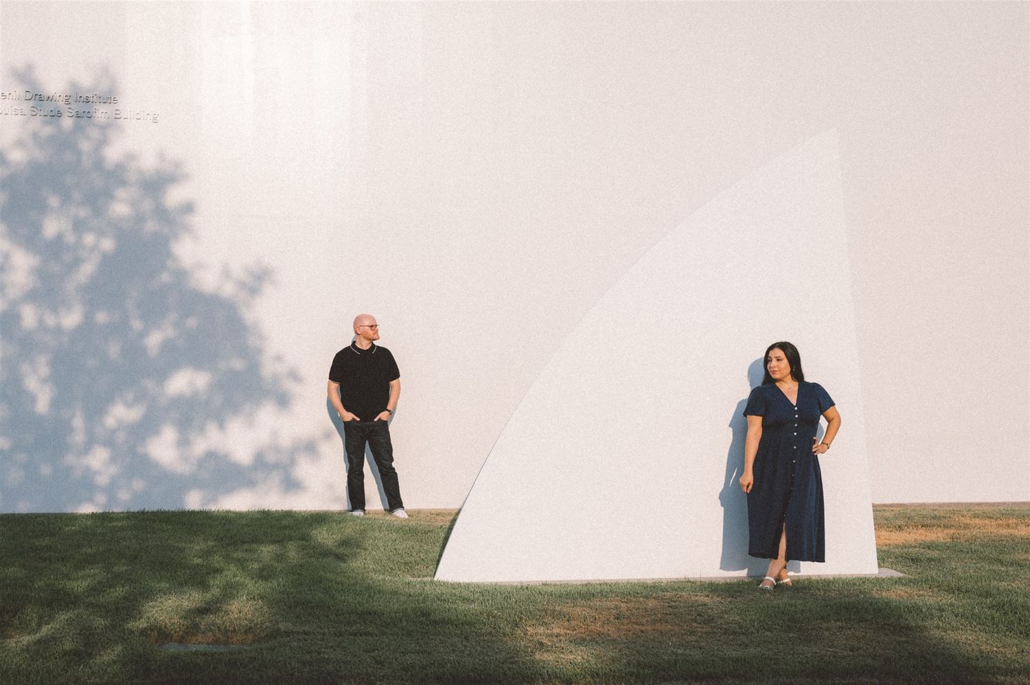 Two figures stand on a grassy hill with a large white architectural structure between them.