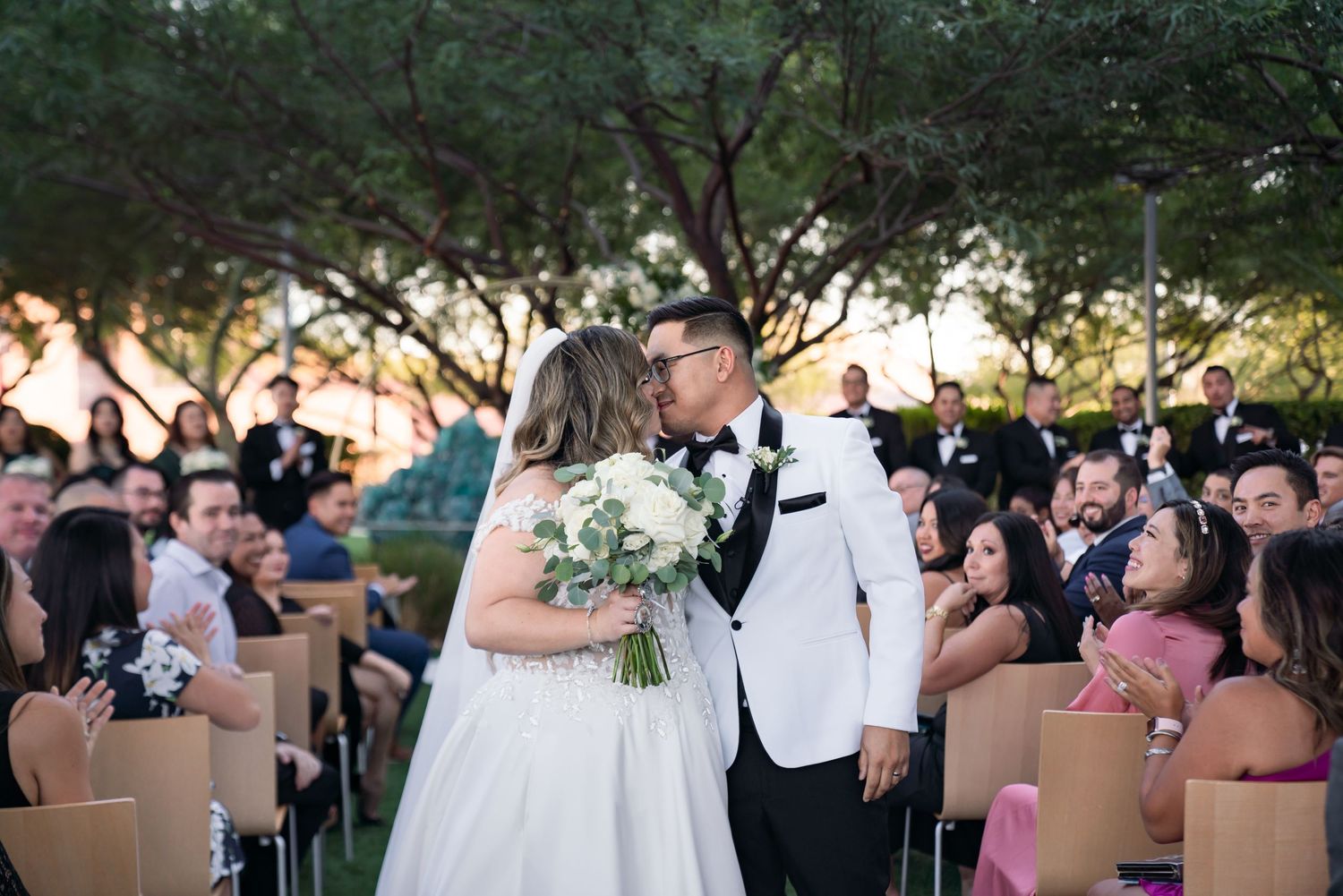 Outdoor wedding ceremony under trees at sunset with guests seated on wooden chairs.
